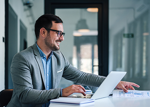 Smiling, relaxed young adult banker at his laptop reaching for papers on his desk.